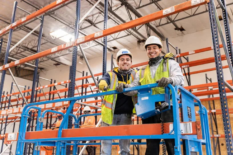 Two warehouse workers smiling under pallet racks, representing responsive customer support and hands-on service.